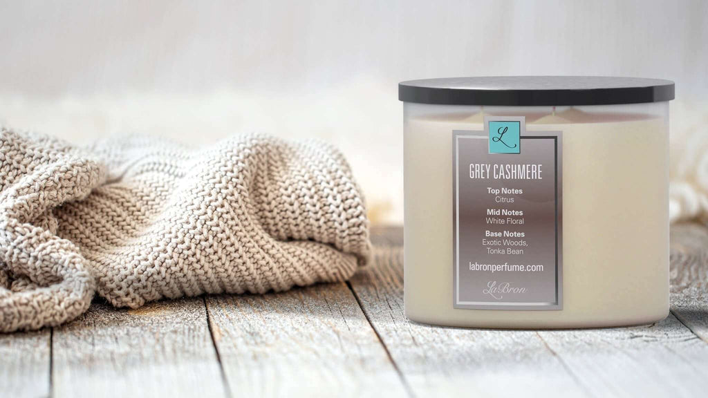 Grey Cashmere candle with a black lid in a warmly lit background with a blanket next to the product. Highly scented with citrus as their top note, white floral with their mid note and finished off with an exotic woods, tonka bean base note.