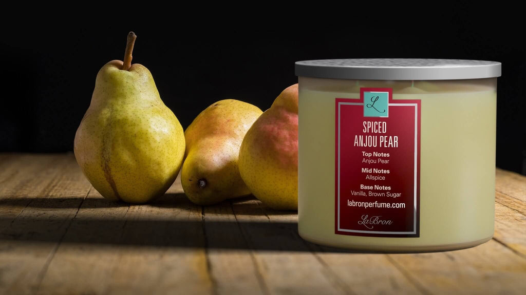 The Spiced Anjou Pear Candle Product next to pears and a wooden floor beneath both objects. This emphasises the smell of the product mixed in with Vanilla and brown sugar base notes.