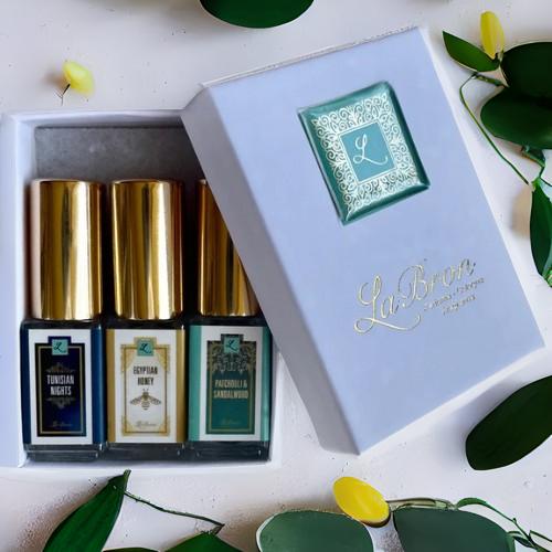 LaBron Perfume Sample Set; with your choice of Three or Six 5 ml Sprays. The box that comes with the products are in a white box with gold lettering for the label. The background is a white background that is surrounded by green leaves.